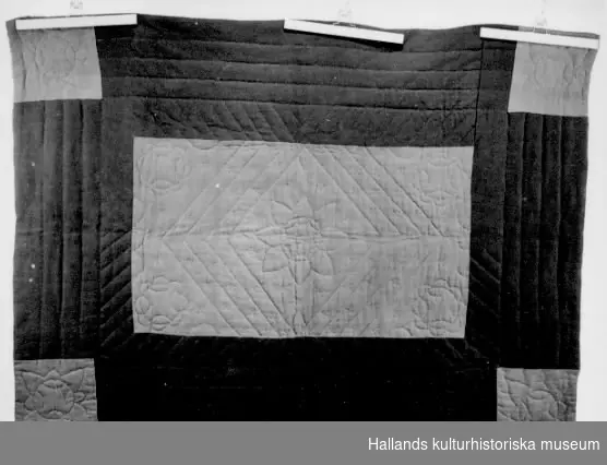 Vaddstickat täcke, genomsytt för att hålla vadden på plats. Sytt som mönster i hörnen och på mitten. Två sidor: översidan sammanfogad av stora tygstycken i rött ( yllekypert) och grönt (tuskaftad vadmal). Undersidan av tuskaftat linne, blå- och vit-rutigt. Påsydd lapp med text "1 st täcke, från 1750, hemspunnet och hemvävt. Martina Andersson, Bjerrome no 5, Vessigebro". Lappen är troligen från en hemslöjdsutställning.