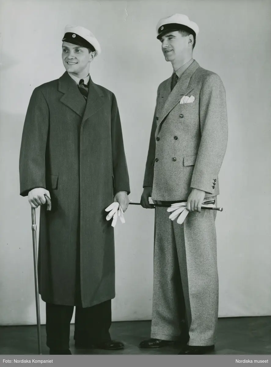 Studentmode 1935. Två unga män i studentmössor och med studentkäppar. Den ene bär en ytterrock med dold knäppning, den andre en ljus, dubbelknäppt kostym. Nordiska Kompaniet. Text med blyerts på baksidan: "MH 1935"