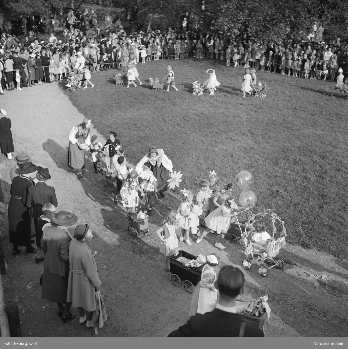 Skansen Barnens dag 1938. Bild över festplatsen tagit en bit uppifrån. Några människor i förgrunden vända mot en blomsterprydd vagn i centrum av bilden. En gräsmatta med fler åskådare i bildens bakgrund.