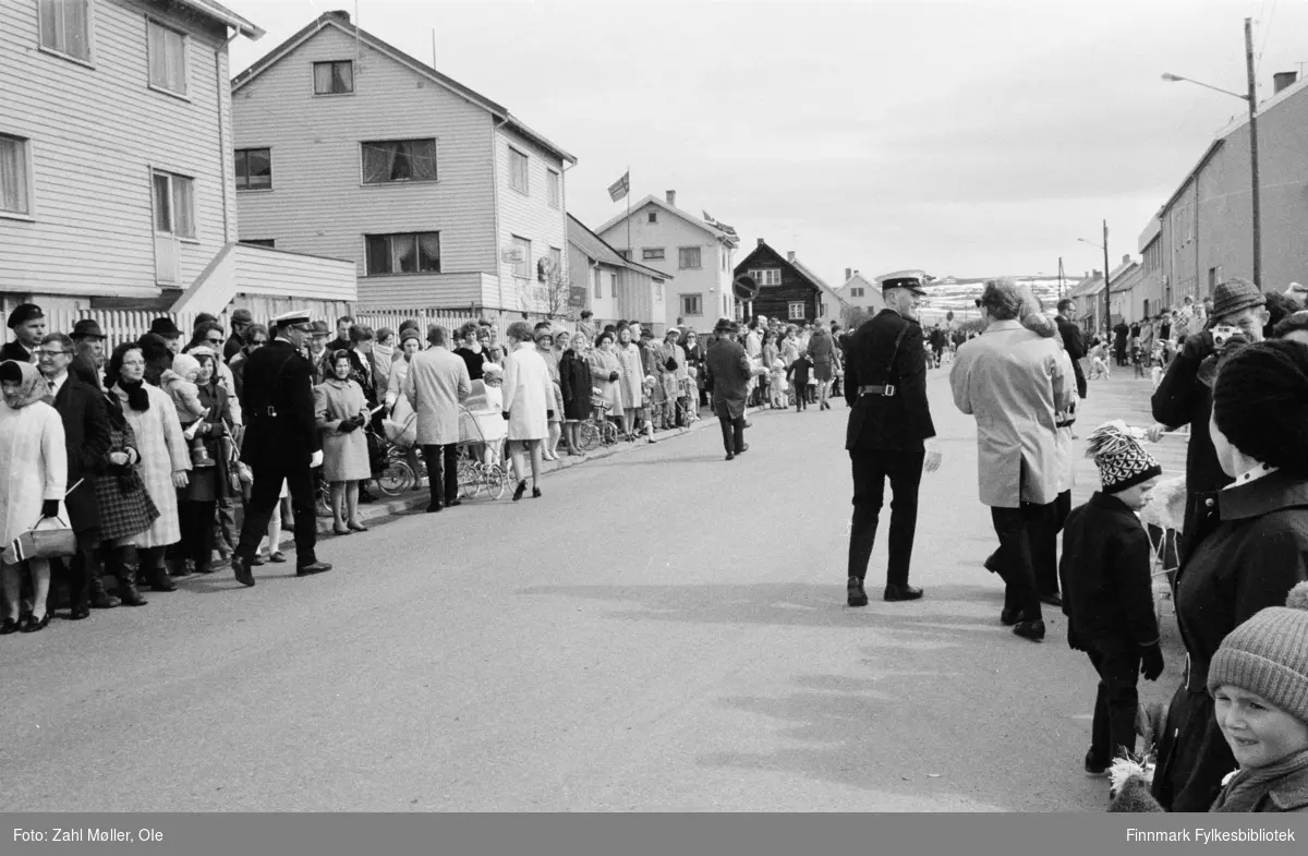 Vadsø 17.mai 1970. Fotografert av Vadsøfotografen Ole Zahl Mölö. Overblikk over alle som står langs gata og venter på 17.mai toget eller kanskje russetoget. Her spaserer politiet til høyre i gata. Ungene bærer luer og de voksne er kledd i kåper og frakker, kvinnene bærer skaut eller hatt på hodet. Flere mødre og fedre med barnevogn har stilt seg på fortauet. Helt til høyre i bildet står en skøyer med kamera og ser rett på fotografen.