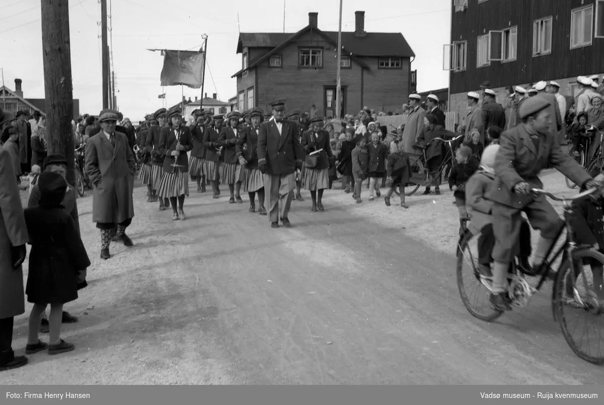 17. maitog i Vadsø 1951. Vadsø jentekorps masjerer, dirigent Olav Holmen går først bak en korpsfane, båret av Vadsø damekor. Bildet er tatt i Nyborgveien, med Alders hvile i høyre hjørne.  Tilskuere på begge sider av veien, antakelig er det Vadsø mannskor som står framfor gjerdet ved Alders hvile. En gutt sykler med et mindre barn på basjebrettet i forgrunnen av bildet.