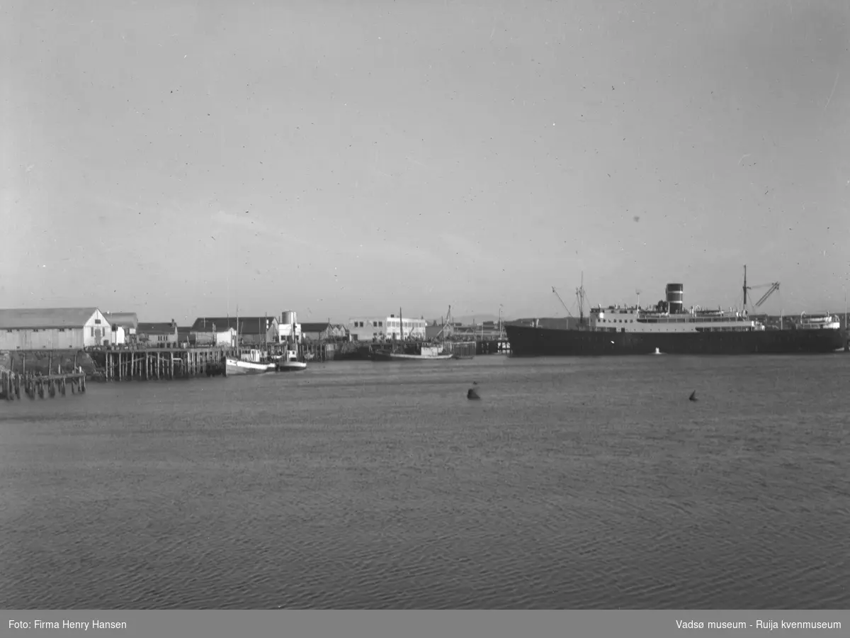 Bildet viser Vadsø havn i 1948 med hurtigruta for anker ved Dampskipskaia. Sentralt i bildet ser en et hvitt hus med flatt tak,  Damskipsekspedisjonen. Til venstre ser en en rekke kaier med tilhørende pakkhus. Tre fiskeskøyter ligger til kai.
