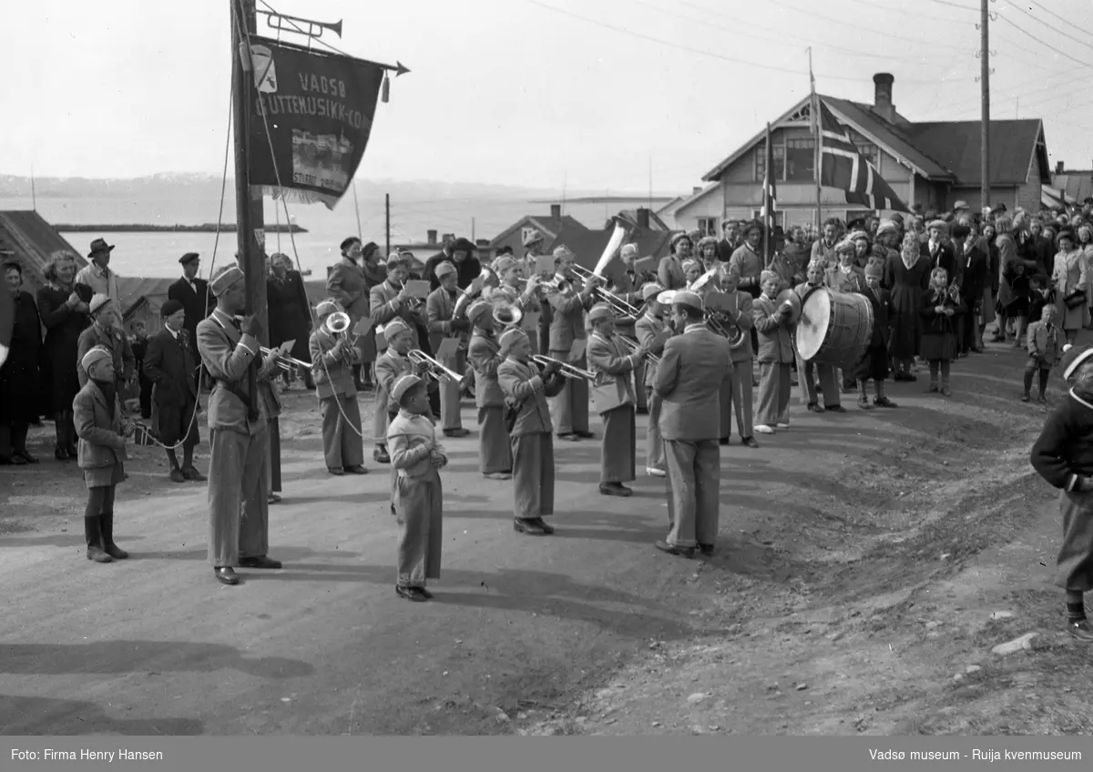 Vadsø 17 mai 1951. 17-mai toget har stoppet i Nyborgveien, foran "Alders Hvile". Vadsø Guttemusikkorps spiller og vi ser en fanebærer med fane. Bakerst i venstre billedkant ser vi havnen og deler av moloen. Publikum, både barn og voksne sees langs 17. maitoget.