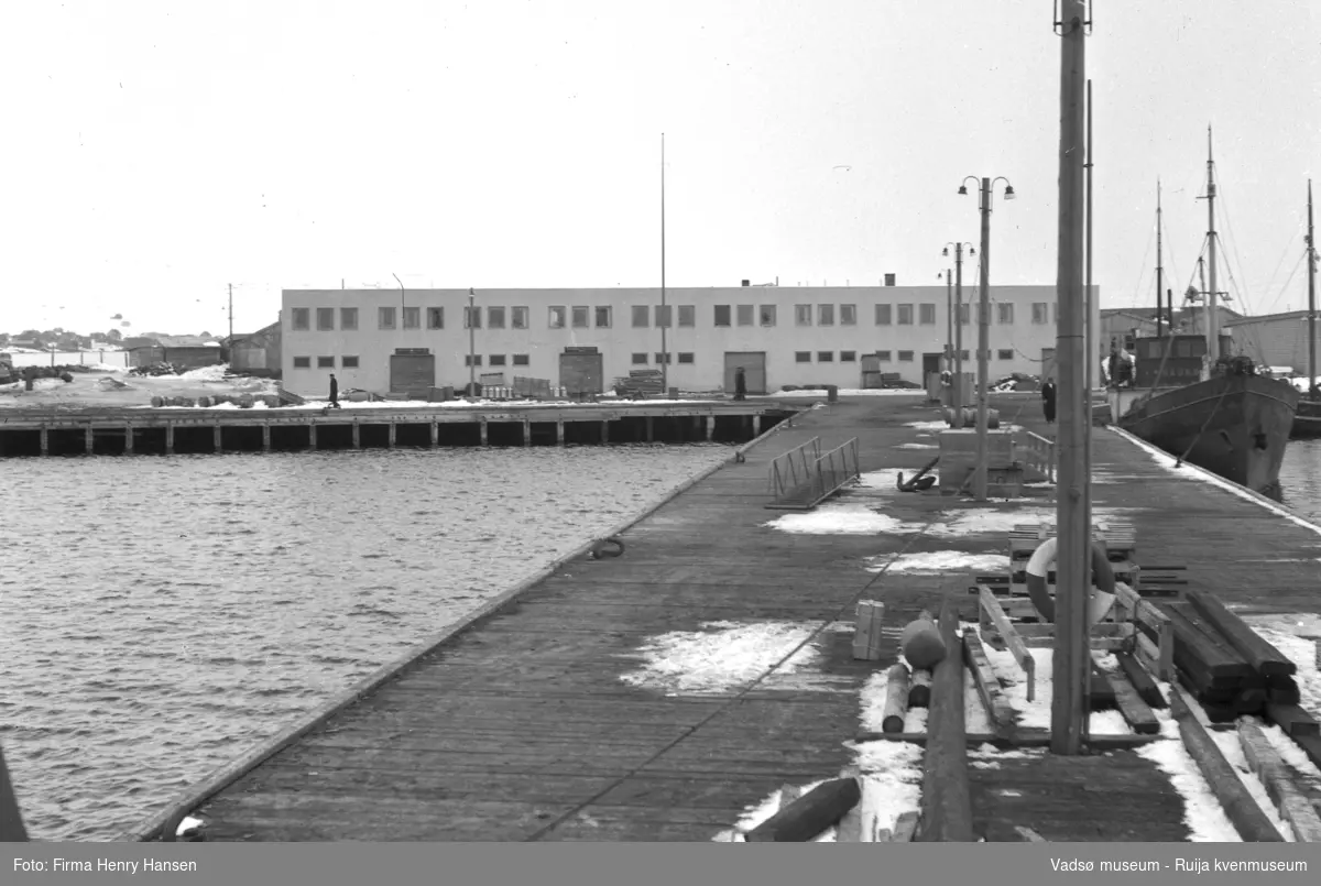 Vadsø våren 1952. Bildet er tatt ytterst fra piren til dampskispkaia fra vest mot øst. I forkant av bildet ser vi piren med blant annet lysstolper, et anker, paller, plankestabler, redningsbøyer og landgangsrampen til hurtigruta. I høyre billedkant ligger lokalbåten Haukøy som gikk i rute på Varangeren for Finnmark Fylkesrederi. I bakkkant Vadsø Havneterminal.