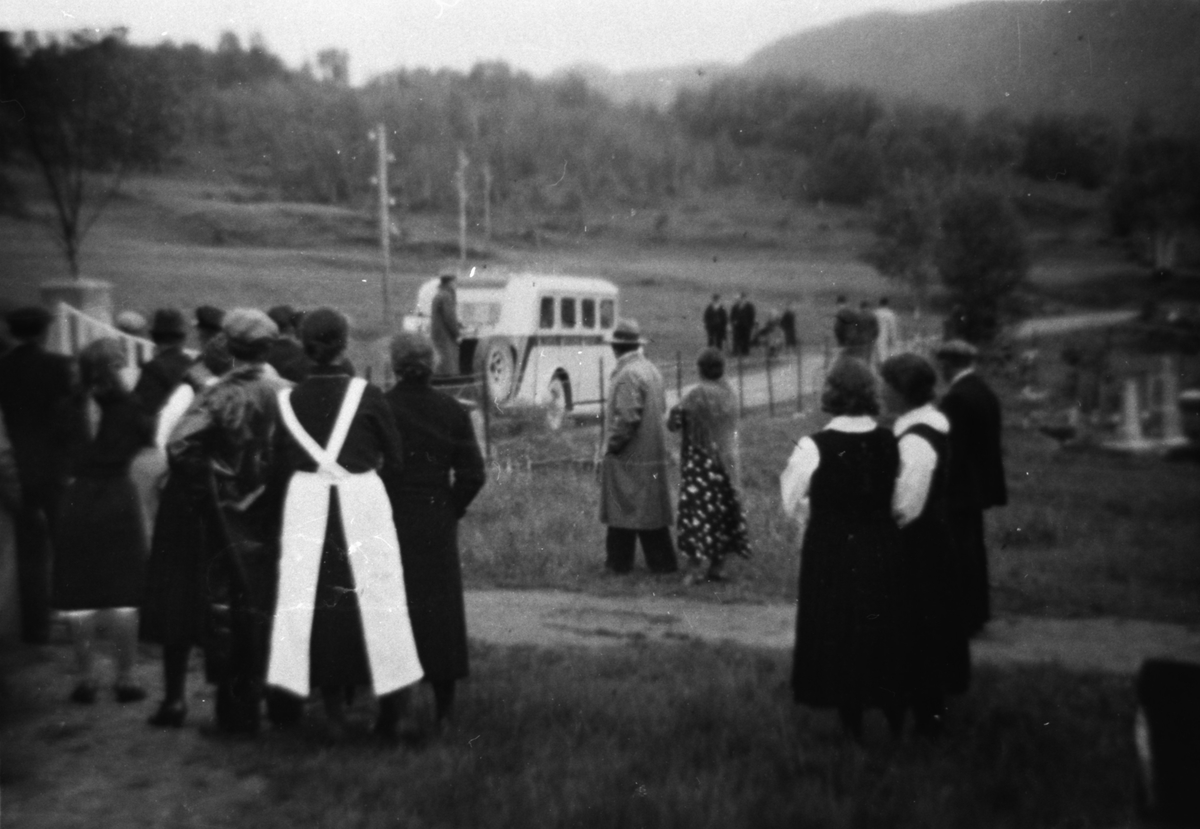 En gruppe mennesker står å ser mot en buss på veien lenger borte. Menneskene ser ut til å være på vei ut en kirkegårds-port.