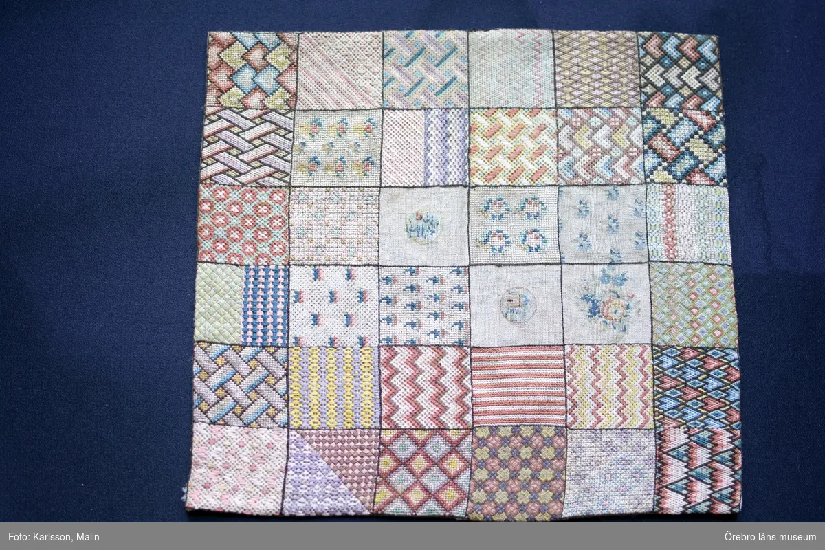 Broderad märkduk bestående av 36 kvadrater med olika mönster.