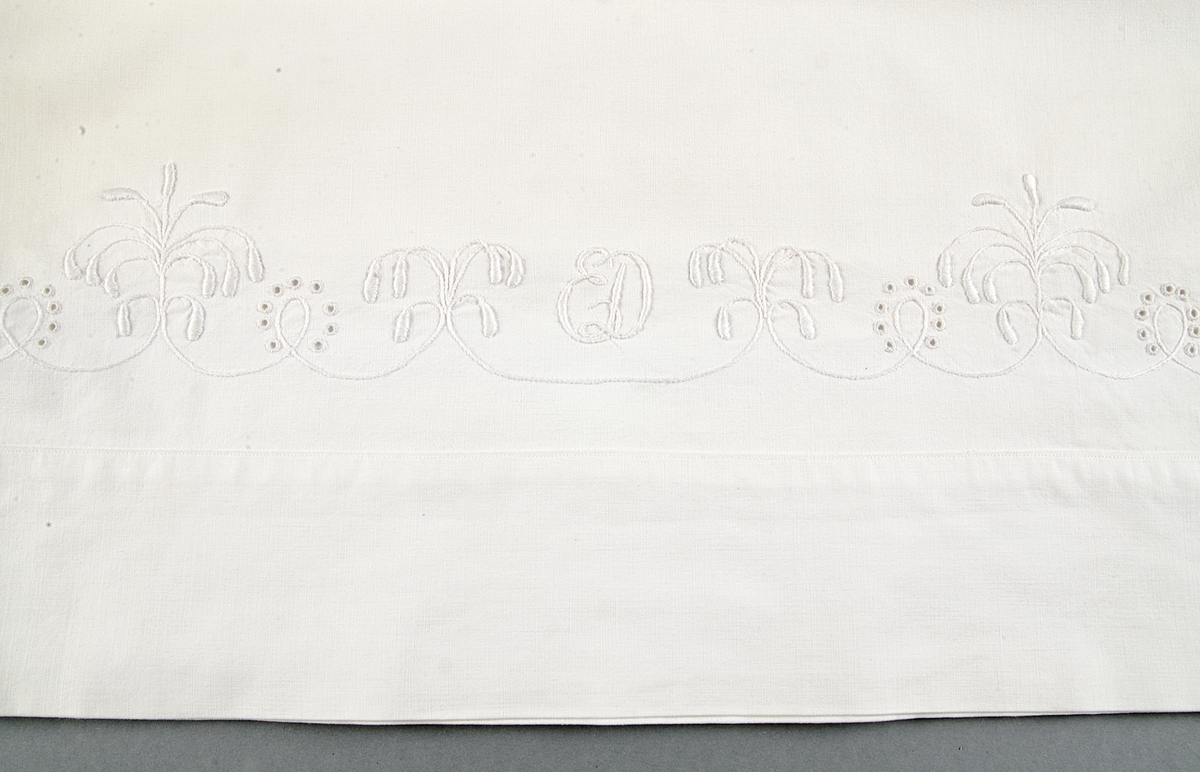 Överlakan av vit bomullslärft, maskinfållat med vitbroderi i bård längs överkanten i klumpsöm, stjälksöm och hålsöm (engelskt broderi), monogram ED.