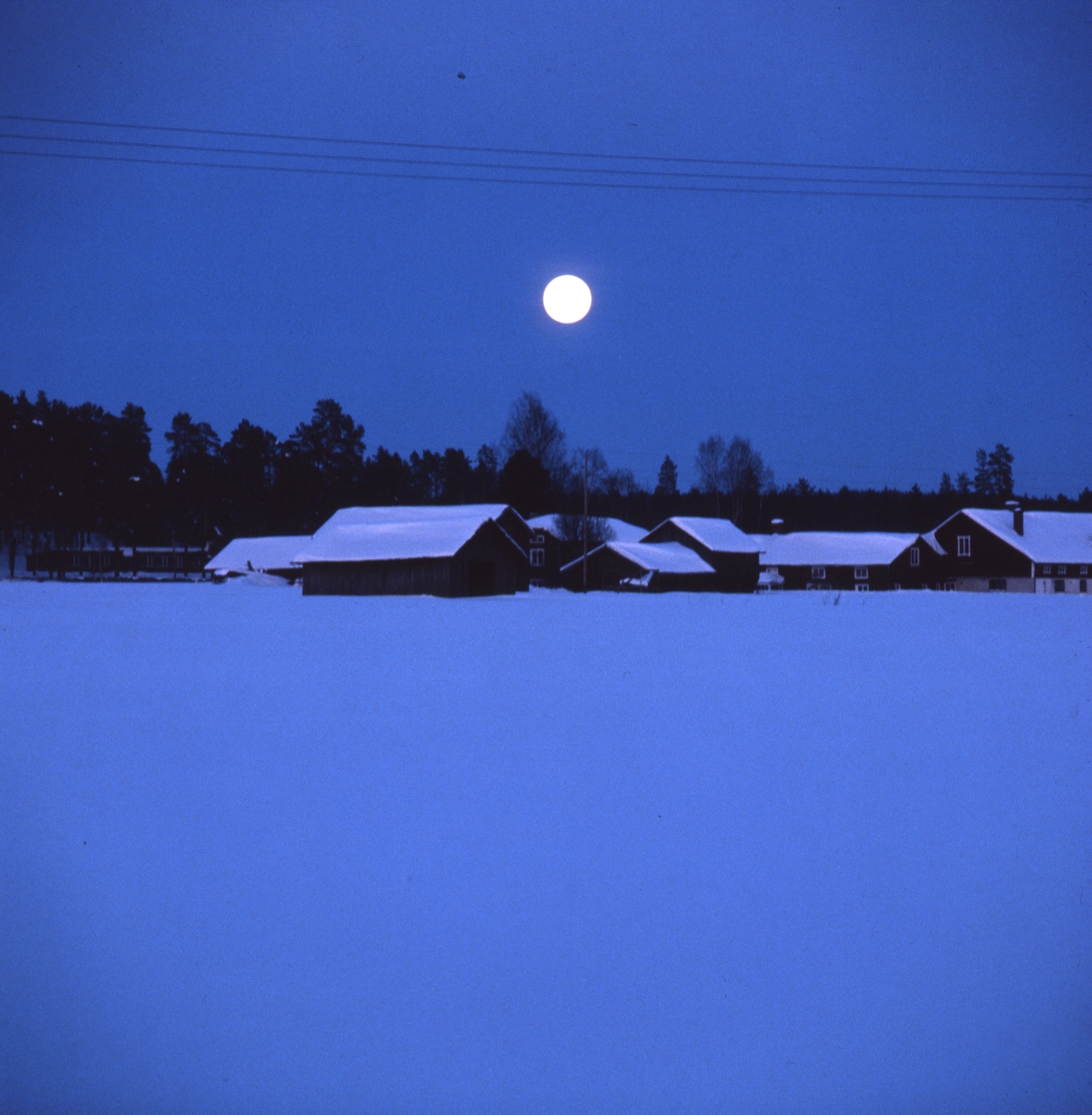 Fullmåne över hälsingegård i Glösbo, Rengsjö. Det är blå skymning och månen lyser på snön på taken.