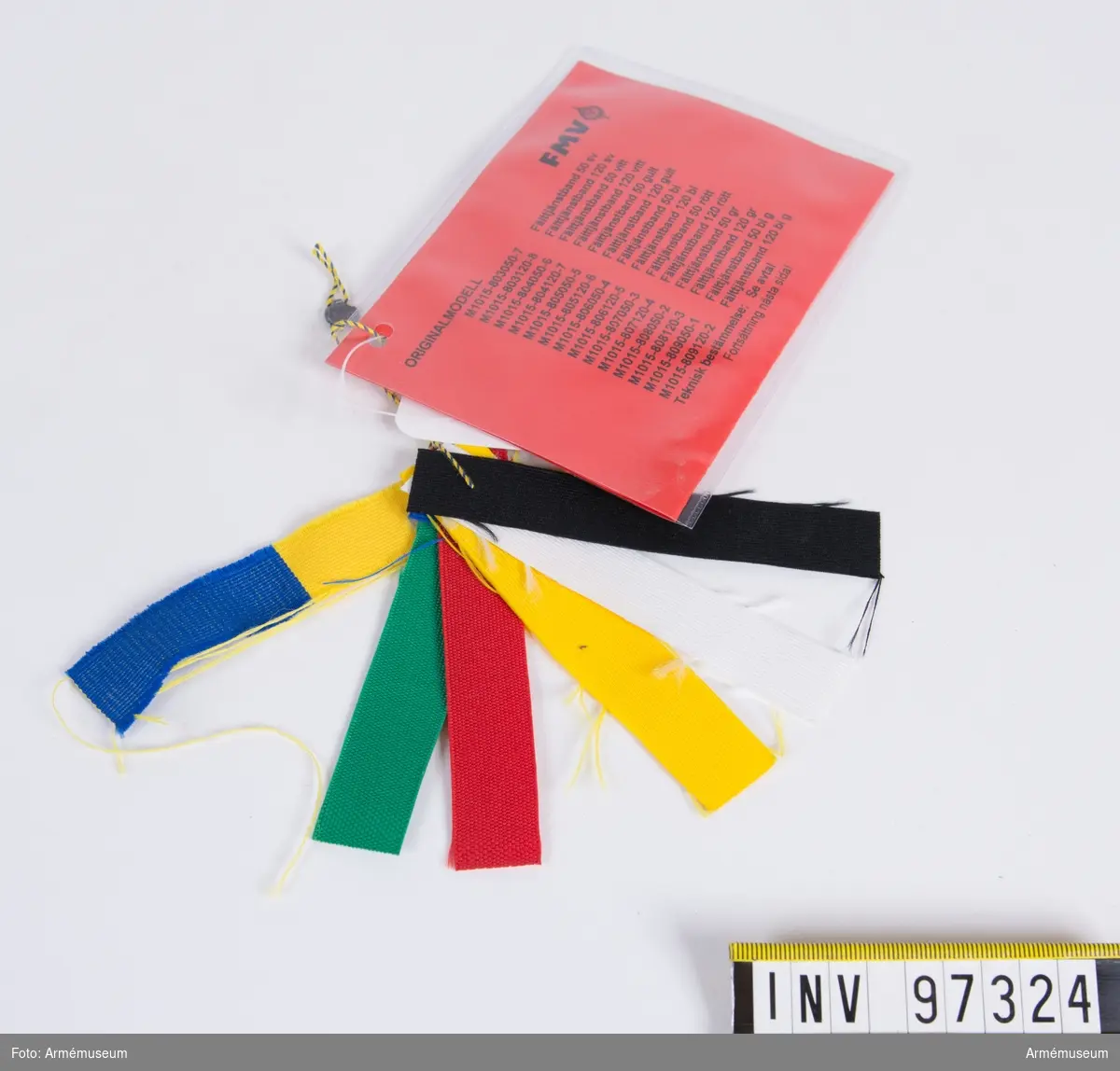 Knippe med prov på band i olika färger. Varje färg har eget M-nummer. På den vidhängande modellappen anges alla M-nummer samt att teknisk bestämmelse finns i avtalet. Lappen är underskriven 1999-01-14 av Roger Strömstedt.