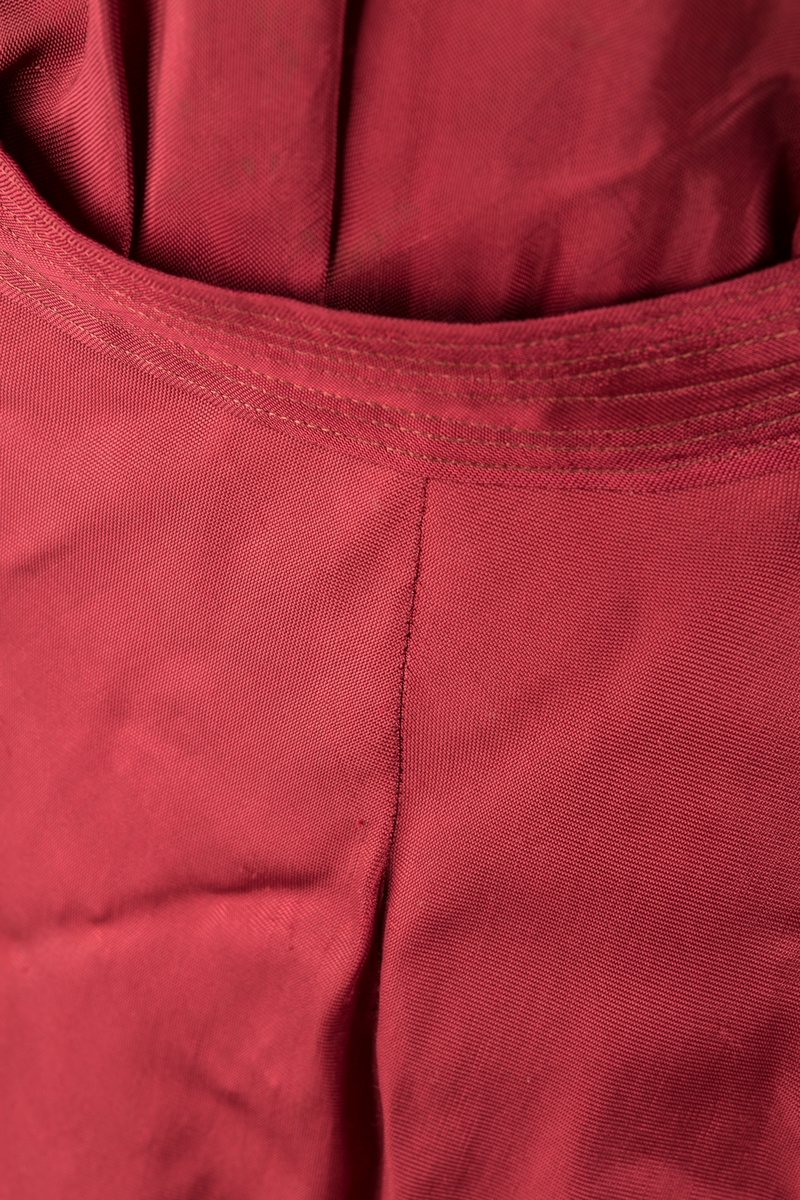 Form: Glatt, klokkeformet skjørt. Forstykkene rynket til buede bærestykker foran. Overdel rynket til skjørt. Korte ermer. Lukket med trykknapper. Belte fastsydd.