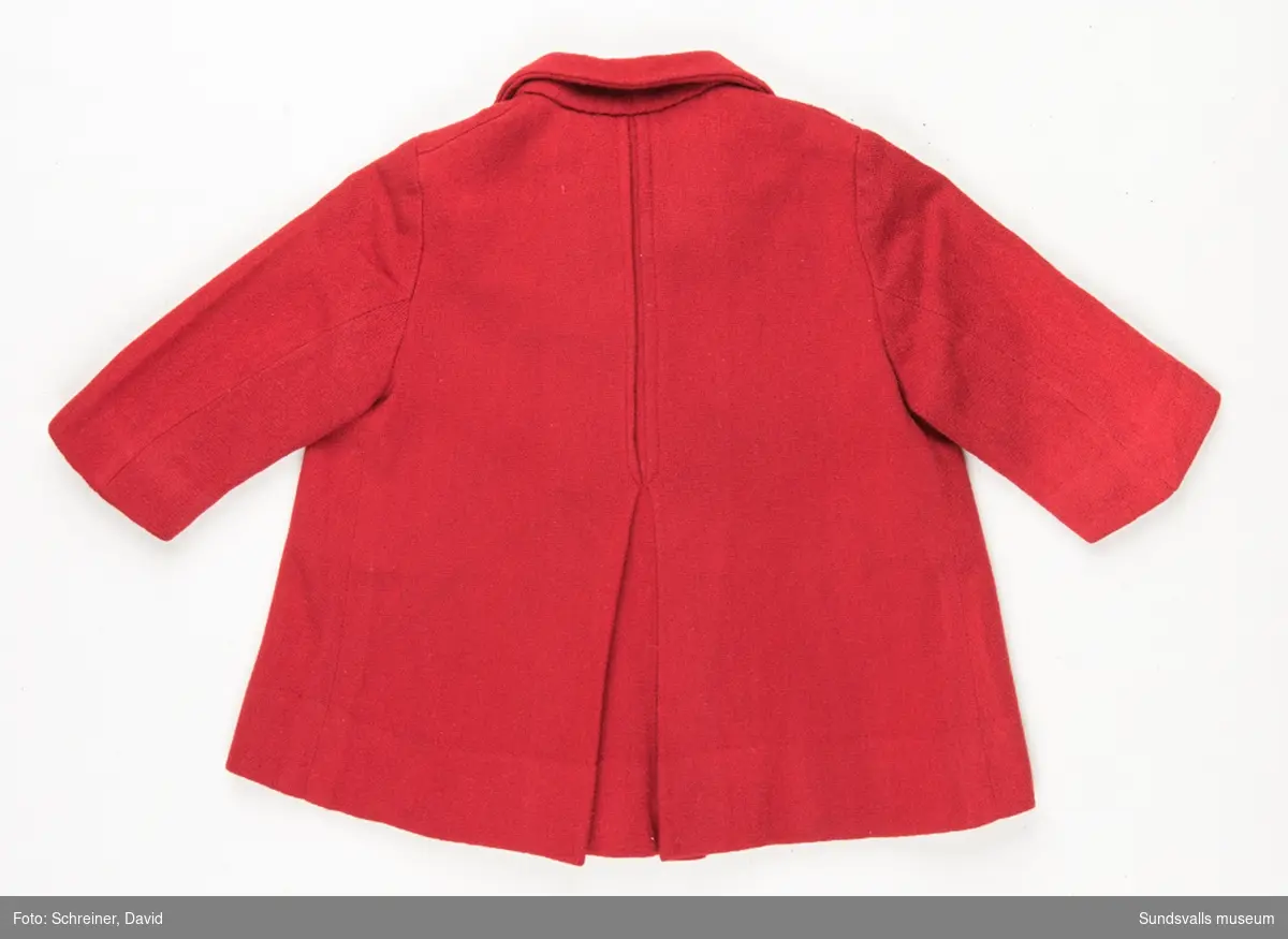 En röd A- formad kappa till flicka i 3-års åldern. Kappan har en rundad krage, dekorsöm och bälgveck baktill. Den övre halvan knäpps med fyra röda plast knappar. Kappan har ett smårutigt tunt bomullsfoder.