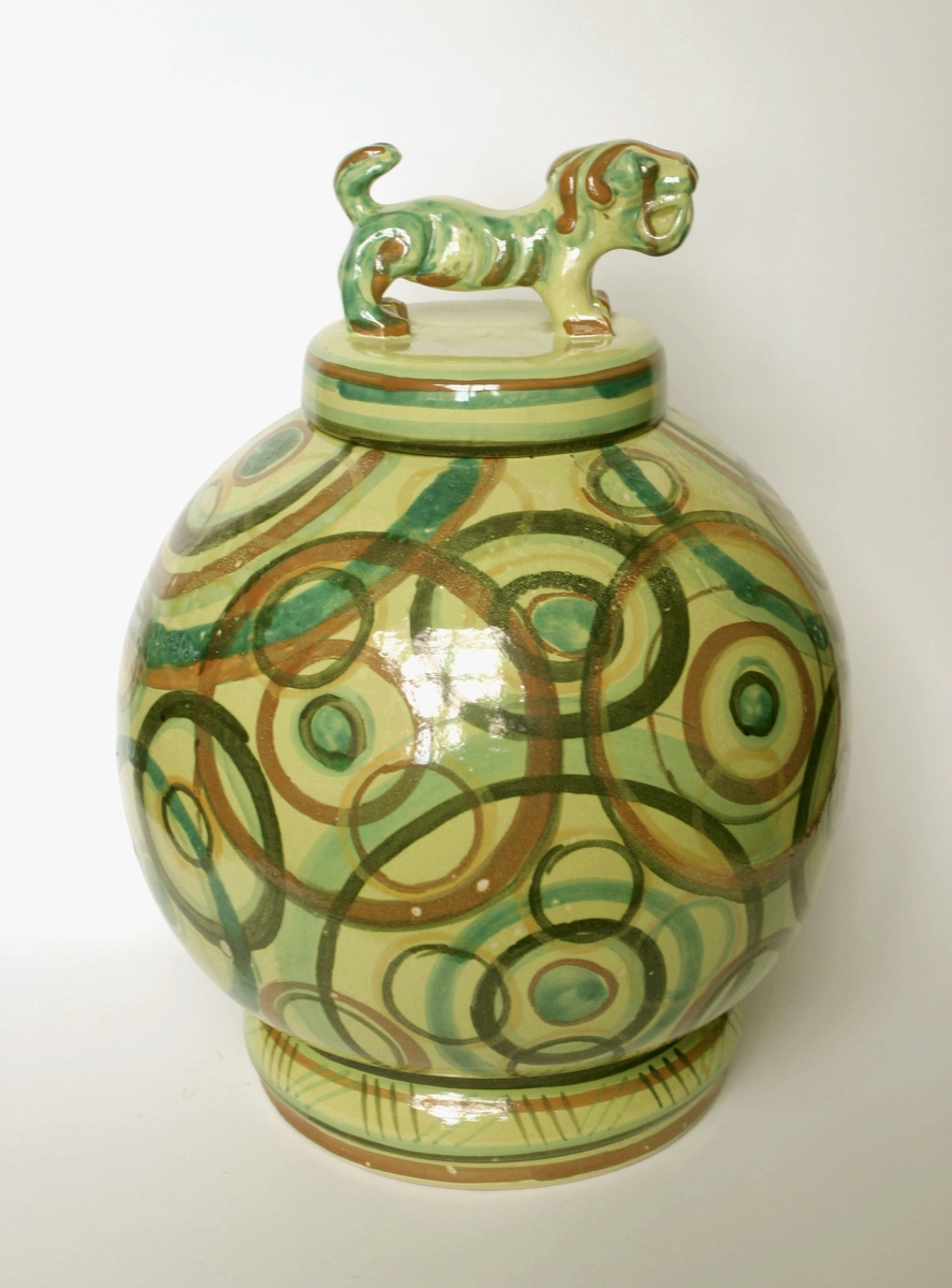 Bojan eller urna med lock, cirkeldekor på grönbrun glasyr på gul botten. Lock med djurfigur/tempelhund. TIllverkare Bo Fajans, formgiven av Allan Ebeling.