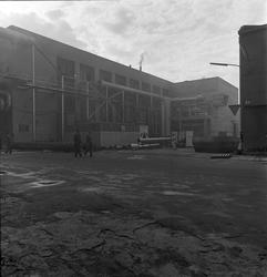 To menn går over plassen ved industribygg.