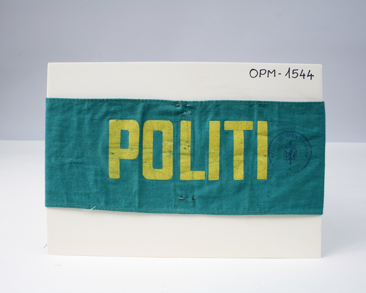 Armbind i grønn tekstil påtrykt tekst "POLITI" i gult.
