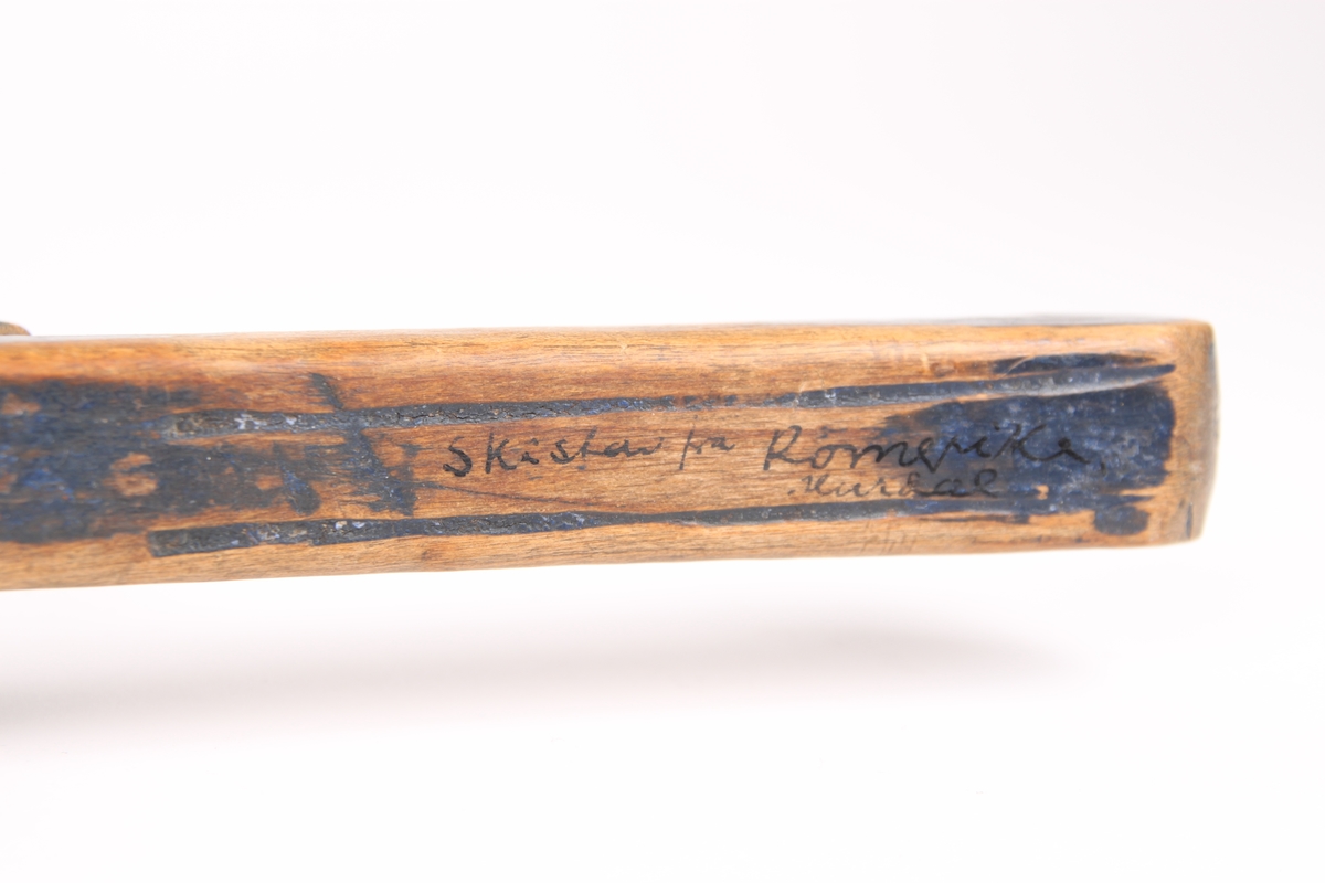 En firkantet skistav i tre. Øverste del av staven har et utskåret håndtak. Navnet til eieren Helge Refsum er skrevet på håndtaket. Nederst på tuppen av staven er det slått inn en liten metallstift.
