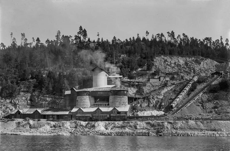 Svart-hvitt bilde av kalkbrenneri sett fra Mjøsa: store, runde kalkovner med tak og kalkbrudd i bakgrunnen.