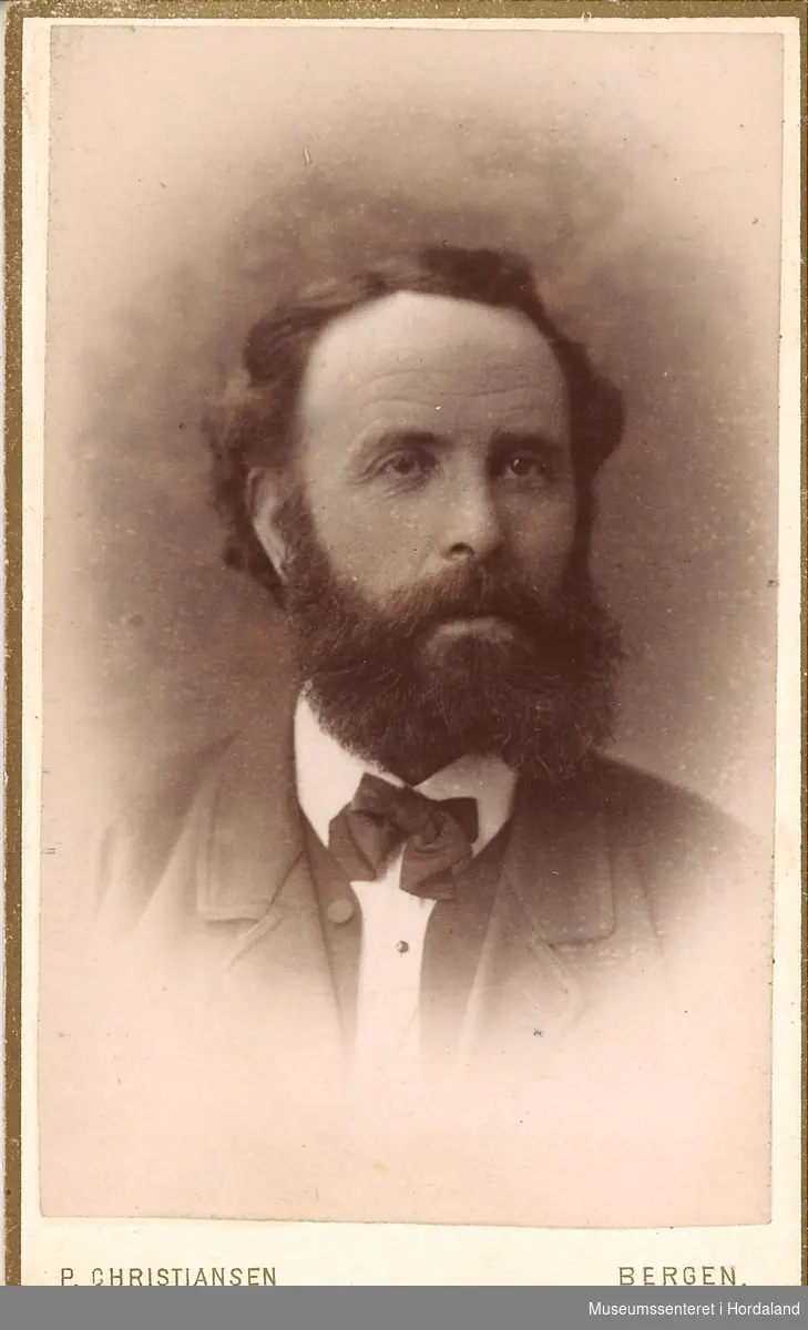 portrettfotografi av mann med heilskjegg, mørk jakke/vest og kvit skjorte med svart sløyfe