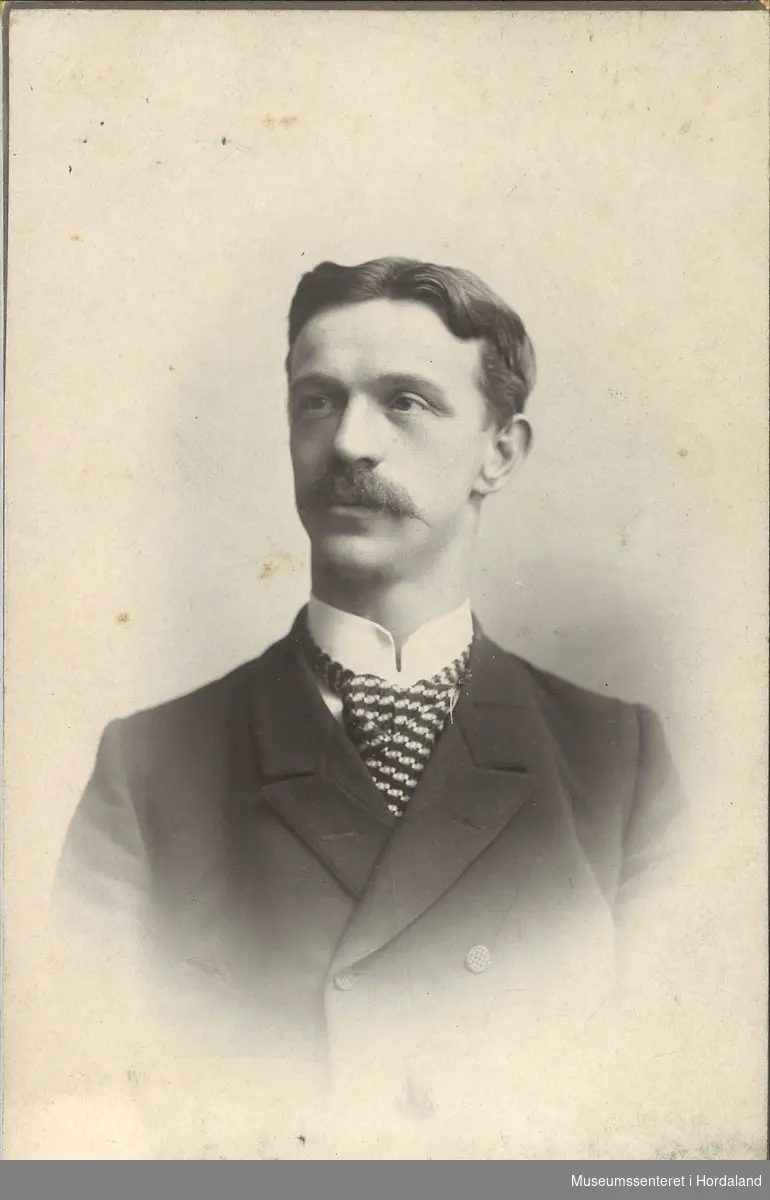 portrettfotografi av ung mann med bart, mørk jakke, kvit skjorte med stivesnipp og stort mønstra slips
