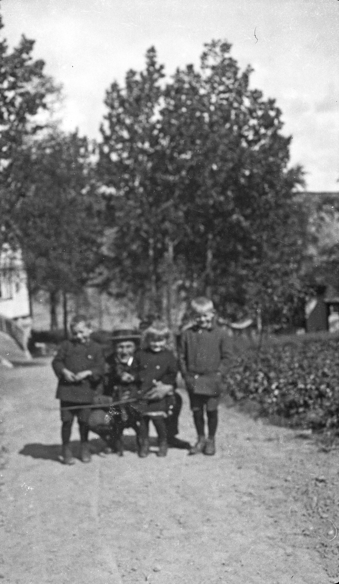 Sommer. Gruppebilde. En mann og tre unge gutter på en grusvei, mannen sitter på huk. Åker til høyre. Skog og fjell i bakgrunnen. Hverdagsfoto.