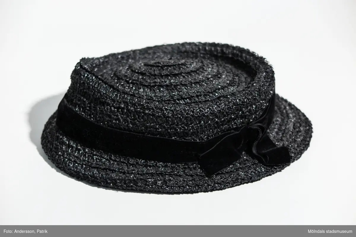 Damhatt inköpt i Eskilstuna, troligtvis på 1930-talet.
Hatten är svart, har ett svart band runt kullen och har en svart gummisnodd som hakband.
Det finns en fastsydd etikett i hatten, men den är mycket sliten och därför oläslig.

Enligt uppgift från givaren är hatten inköpt på 1920-talet, men eftersom butiken öppnade 1934 är hatten troligen inköpt på 30-talet.