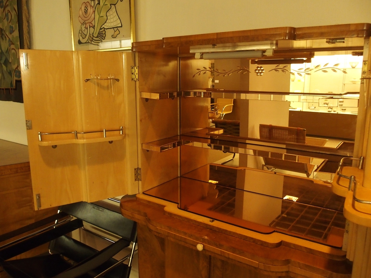 Cockailskap i to høyder med fire dører. Profilert korpus, sokkel, gurtlist og topplist. Mahogny med valnøttfiner; de øvre dørene er finert med lyst satintre på innsiden. Nedre skap har en hylle og en skuff festet øverst. Gurtlisten har en uttrekkbar hylle med ferskenfarget speilglass. Øvre del av korpus har dører som åpner til skap med belysning. Veggen bak, bunnen og to hyller er dekket med ferskenfarget speilglass. Øverst på veggen bak ses geometrisk dekor flankert av bladranker. På hver av dørenes innsider en hylle og gjennomhullet feste for cocktailpinner. Ti originale glasspinner med svaner er bevart.