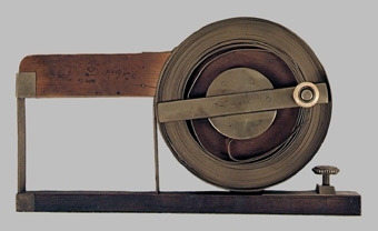 Måttband av mässing. 26 alnar långt. Tillverkat av Elias Nyman. Handtag bakom en spole med vev som matar fram och tillbaka måttbandet.