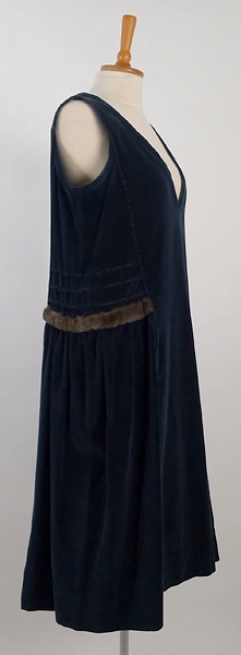 Klänning, av blå sammet med moaré effekt. I vardera sida finns tre sydda veck och under dem en skinngarnering. Klänningen är delvis handsydd. Vid användning en blus under.