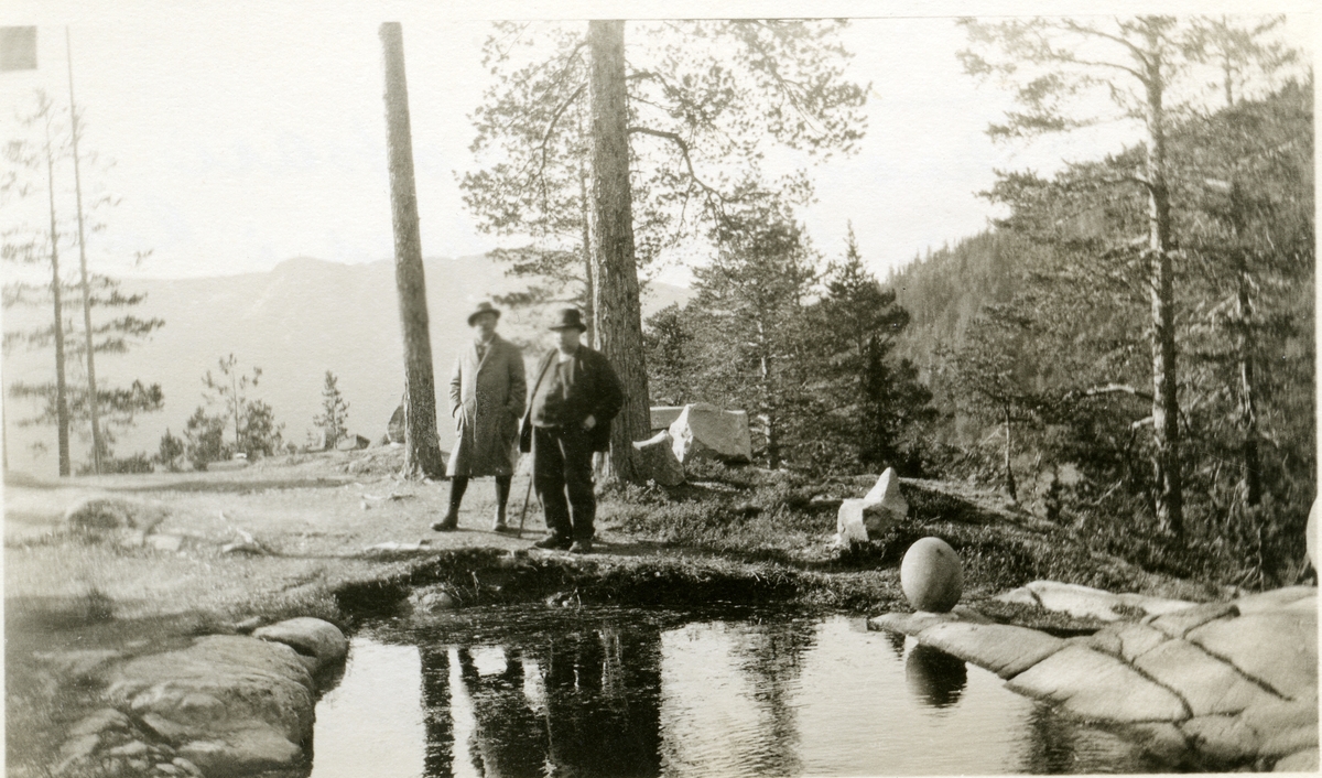 2 menn står ved en dam, med fjell og skog i bakgrunnen. Vatneberget. Den ene har stokk.