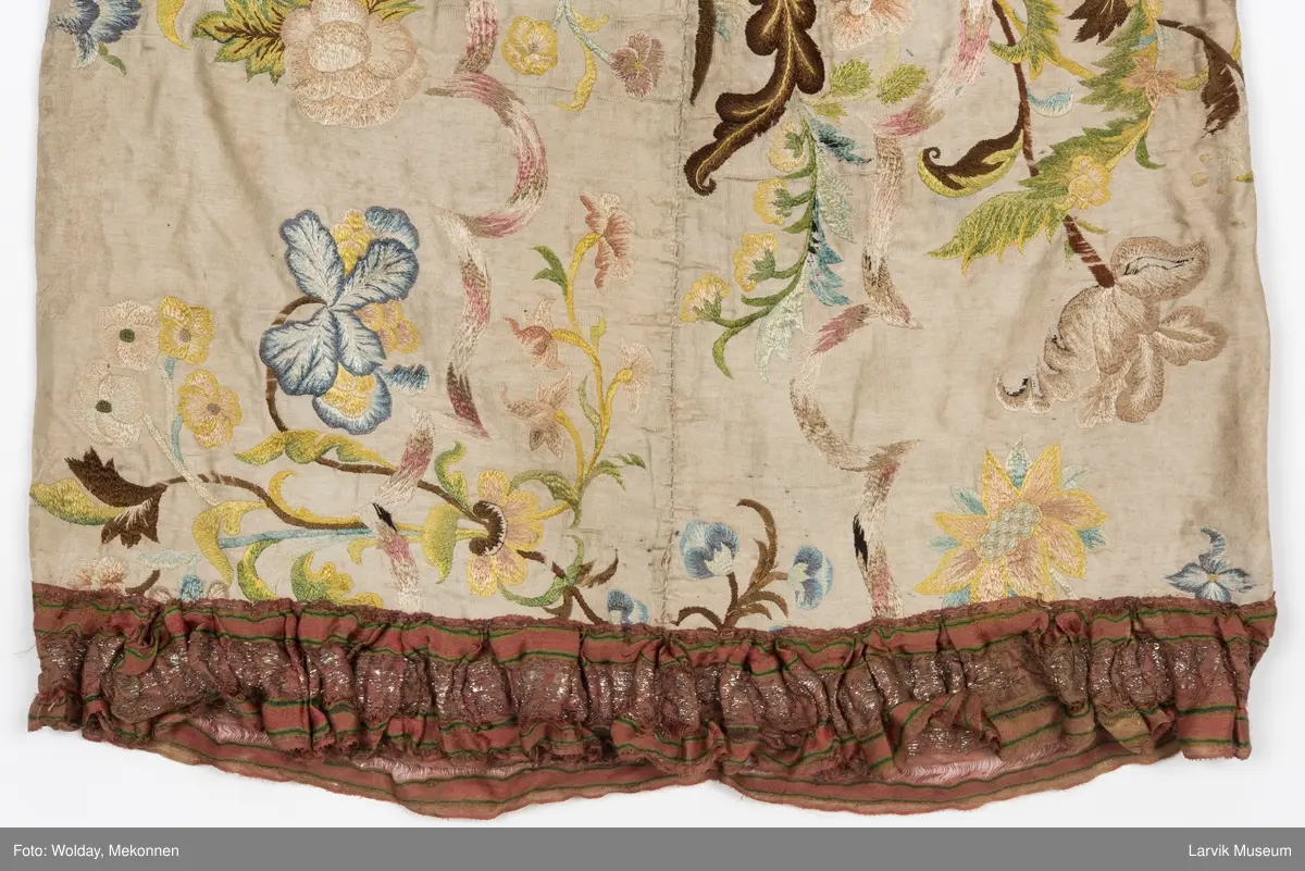 Form: livstykke som hette, åpen til livet, bred linning, rynket skjørt med blondekant nederst, søm bak på hetten i linning, søm foran og bak i skjørt, enkelte skjøtesømmer, linning brodert med gulltråd, skjørtet er foret med tynt stoff