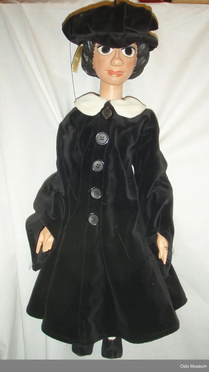 Stangmarionett, dame.
Hodet er laget av gips(?). Ansiktet er malt i en rødlig hudtone. Øynene blå og munnen ferskenfarget. Håret også gips(?), malt sort og tvinnet som en krans rundt hodet.
Marionetten har på seg en sort fløyelshatt, sort fløyelskåpe med sorte, blanke, runde knapper og en hvit fløyelskrage. På bena, sorte fløyels snøresko med brun skinnsåle.
Kroppen er i tre. Det er bevegelige treledd i hofter og knær. Leggene er malt hvite. Armene er bevegelig i skulder-, og albueledd. Skinnremser er spikret på og danner leddene.
Fem sorte tråder er festet til kroppen: på knær, håndledd og rygg. 
På hodet er det festet jernstang med et rødt trehåndtak og en rød bevegelig treform der trådene er festet.