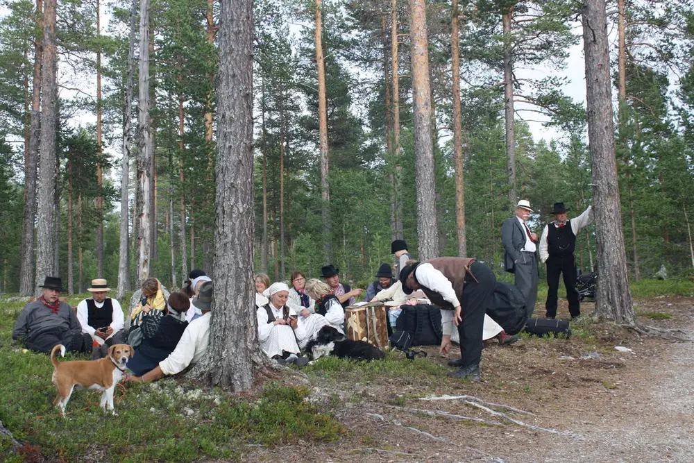 En gruppe mennesker i tradisjonelle klær sitter i skogkanten.