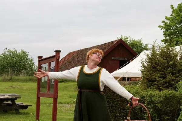 Rødhåret kvinne i middelalderdrakt, med kurv i hånda, slår ut med armene for å ønske velkommen til Urtehagen. Foto/Photo