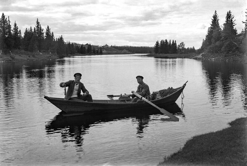 Svart-hvitt foto av to menn i en åfløy (robåt) på et stille vann eller ei elv med granskog på flere sider.