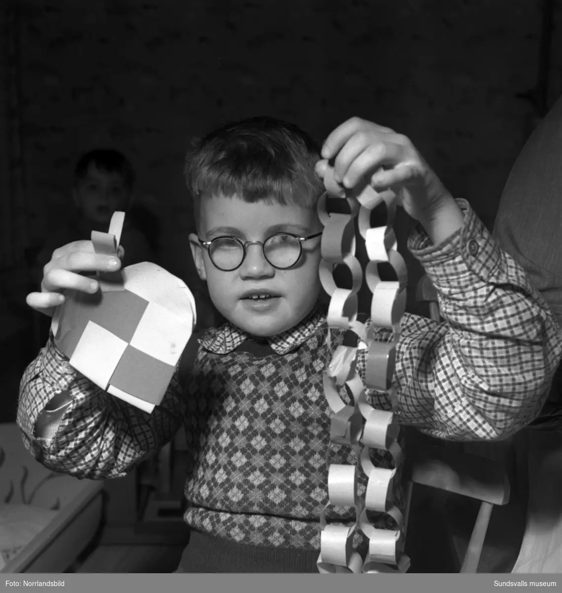 Barn och personal på barnhemmet Morgonsol tillverkar juldekorationer. Branta vägen 8, Södermalm. En liten pojke visar stolt upp en pappersgirlang och ett flätat hjärta.