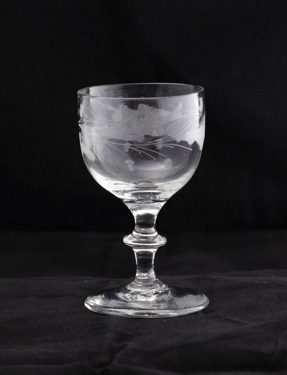Empireglass med rett likesidet rundbundet klokke, sylindrisk stett med en skarp knapp og flat fot. Glasset er klart og er dekorert med ekeløvfrise gravert.