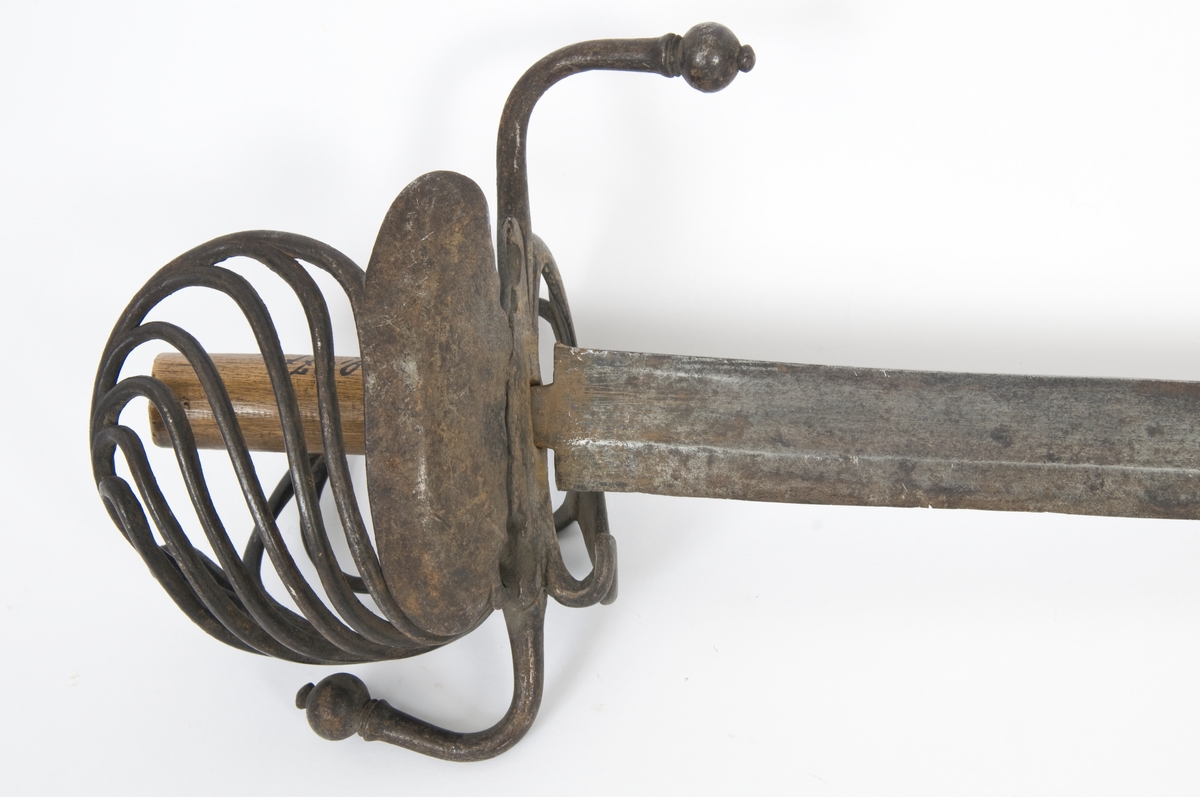 Knapp mangler. Tregrep. Asymmetrisk feste med runde bøyler. Fremre parerplate er nyreformet, og innsiden har tombøyle og klingebrekker. S-formede parerbøyler som ender i en kule.  
Enegget klinge med bred blodrand og ryggslipning mot spissen.  Gjeddenebb.  Smedmerke på innsiden.
Balg medfølger.  Trebalg trukket med 2 lag lær. Balgen er i dårlig stand.