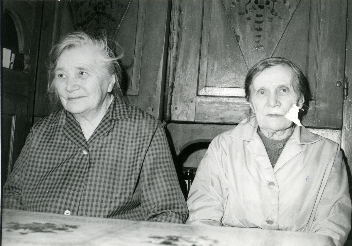 Kari og Jørgine Vestgrøv sitter ved et spisebord. De har et gammelt framskap bak seg.