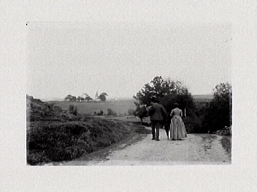 Kvinna och man ses bakifrån promenera på grusväg i öppet landskap med kyrka i fonden. Mannen har ett paraply. Eventuellt Alfshög. 1890-tal.