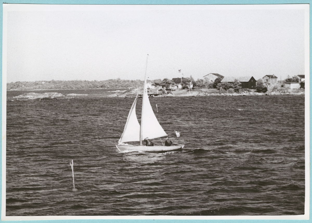 Från kappseglingen. En seglingsjolle med två besättningsmän syns färdas över vattnet framför en av öarna i skärgården, troligtvis Basareholmen.