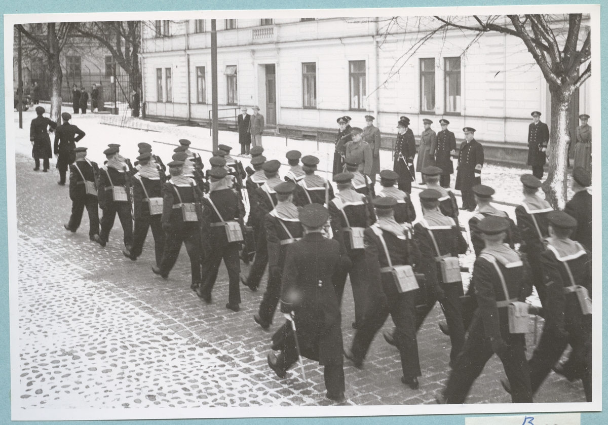 Uniformsklädda flottister marscherar längs med den snötäckta Drottninggatan. Gatan kantas av Sparres kanslibyggnad. Utanför byggnaden står militärer av högre grad.