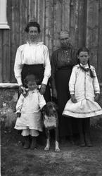 To eldre kvinner, to småjenter og en hund foran en husvegg. 