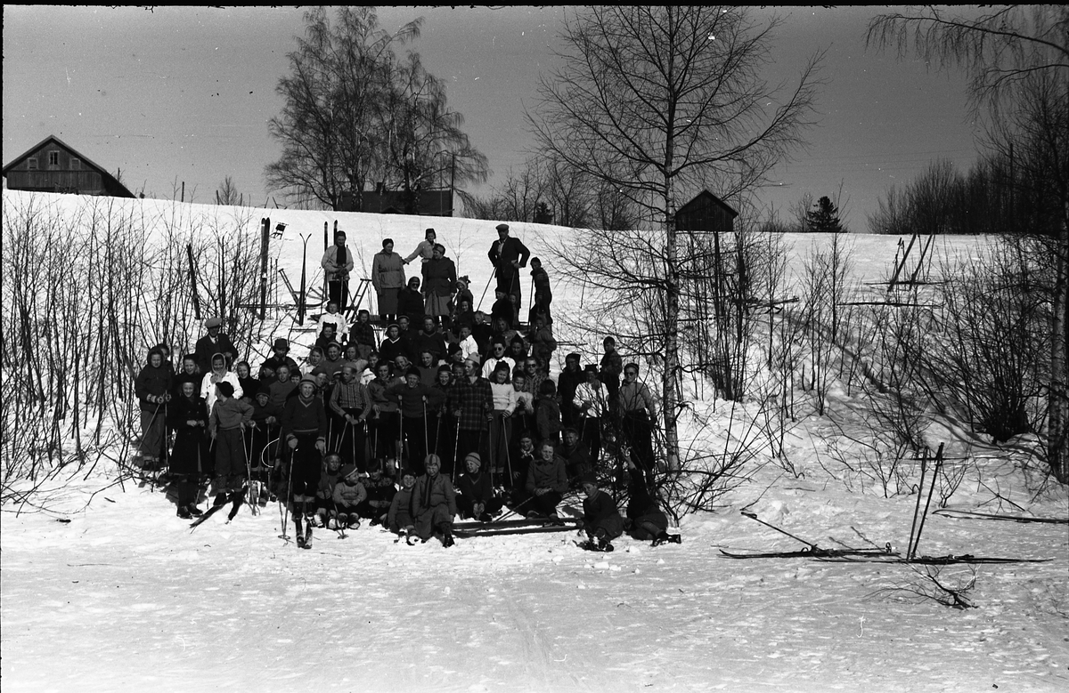 En rekke barn og noen voksne på skitur. Muligens skolebarn fra Hovsvangen på utflukt til Løken i Vestre Toten, og bilde tatt nede ved Slomma. Den høge mannen til høyre bak kan minne om lærer Ole D. Haug. Ingen andre er identifisert. Vinteren 1950.