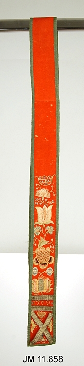List till folkdräkt, Värendsdräkten. Tillverkad av rött ylle som är hopsytt av tre remsor, kantad med grönt tyg, fodrad med linnetyg. Broderiet i flera olika färger av silkesgarn i form av olika blommor, monogram "C S D, 17 9 2" eller "1762" (bakvänd 9a, eller uppochnedvänd 6a?). Dekorerad med metallband som går i kryss i ändarna, ovanför detta två vågräta band.