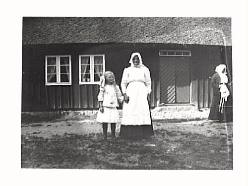 Äldre kvinna håller en flicka i handen. De står mellan ett par fönster och ingången till ett bostadshus med halmtak.  Till höger står ytterligare en gammal kvinna.
