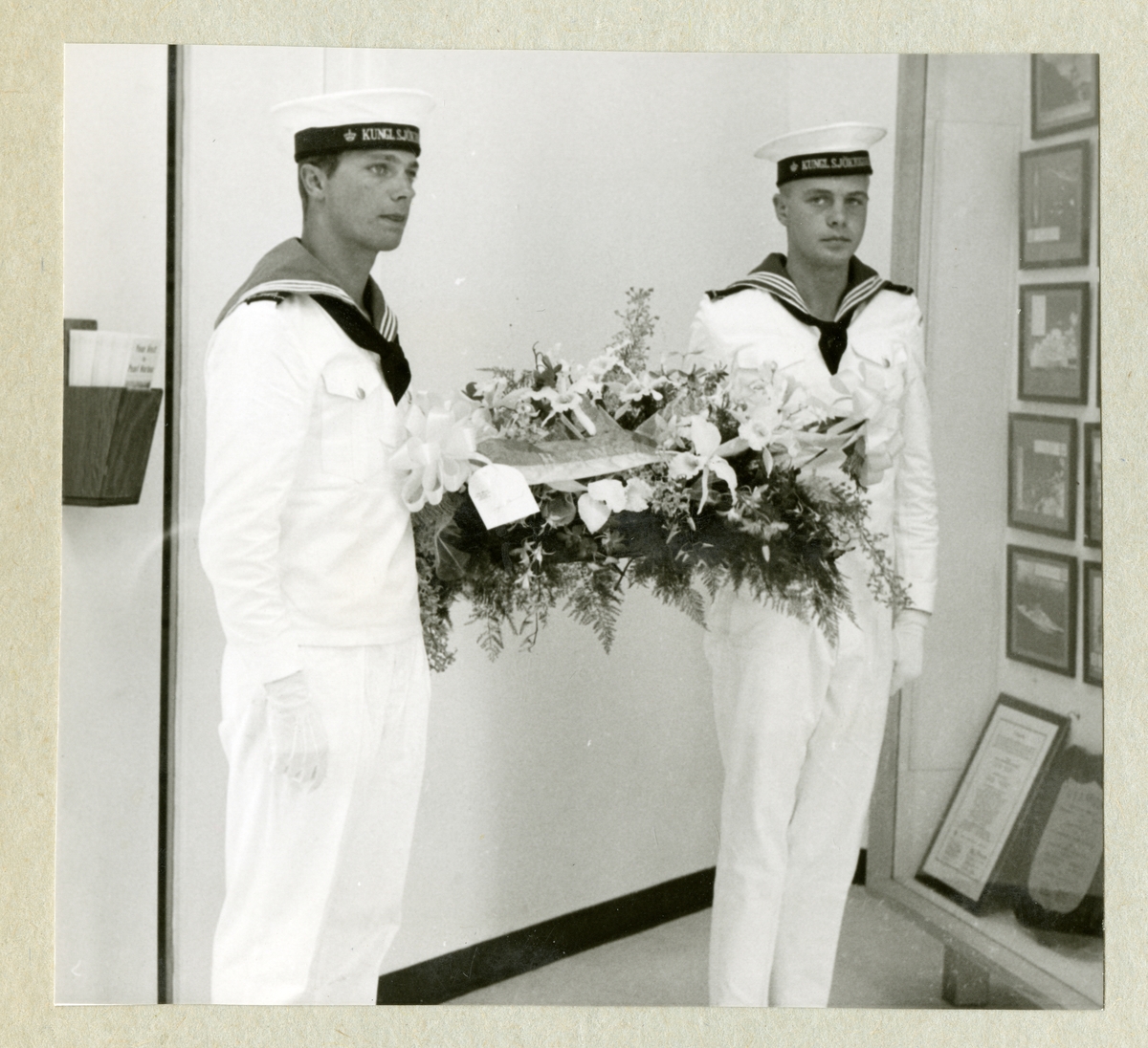 Bilden föreställer en besättningsman och Carl XVI Gustaf i vita uniformer. Mellan sig håller de ett stort blomsterarrangemang. Bilden är tagen under minfartyget Älvsnabbens långresa 1966-1967.
