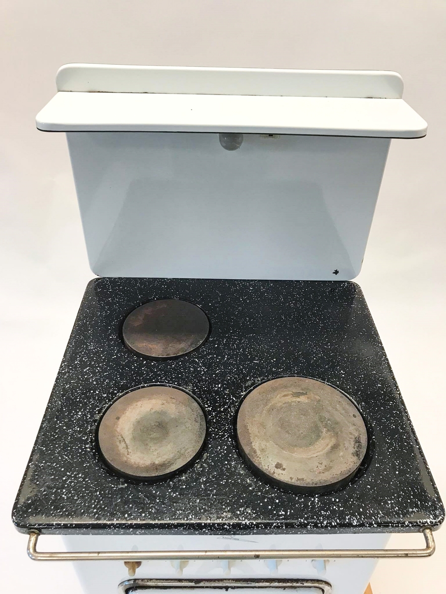 Elektrisk komfyr med bakvegg og tre kokeplater. Stekeovn med termometer på utsiden. I stekeovnen er det to tilhørende stekebrett. Lyspære på bakveggen.