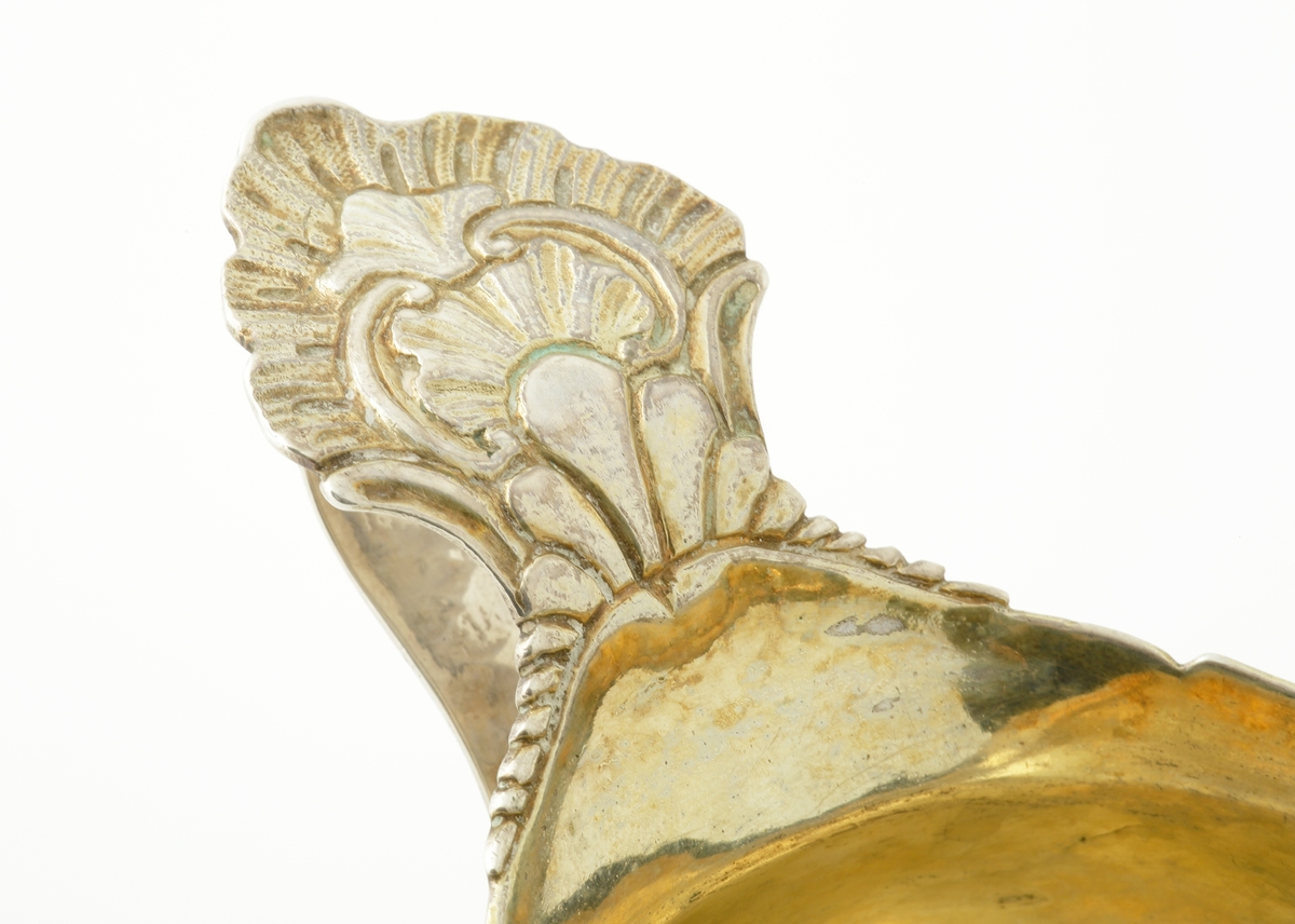 Gräddsnipa i silver. 
Oval , något bukig modell på fyra fötter, dekorerade med blad. Flikig övre kant. Invändigt förgylld. Hänkel med bladdekor. Stämplar på hänkeln.