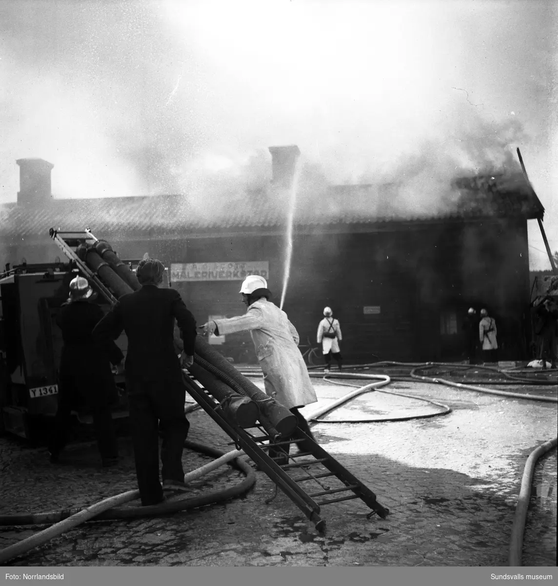 En brand i den gamla vinfabriken som då låg väster om Grönborgs bryggeri. Brandmännen jobbar med att släcka elden som uppstått i den västra delen av byggnaden och även en liten kiosk vid byggnadens västra gavel är hotad. Hela den timrade byggnaden flyttades senare till Norra berget.
