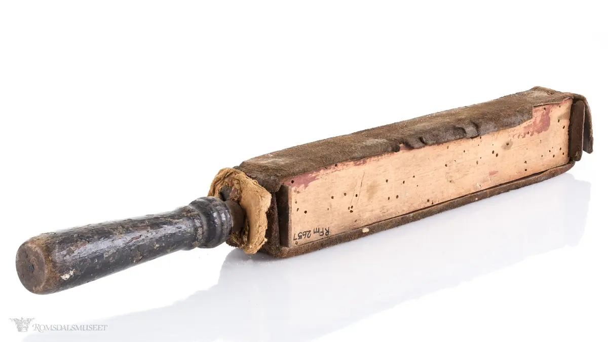 Rektangulær med dreid trehåndtak. Lærrem som går rund hele. Avstivet med metallbeslag foran og bak. Brukt til å stroppe/kvesse barberknivblad før bruk.