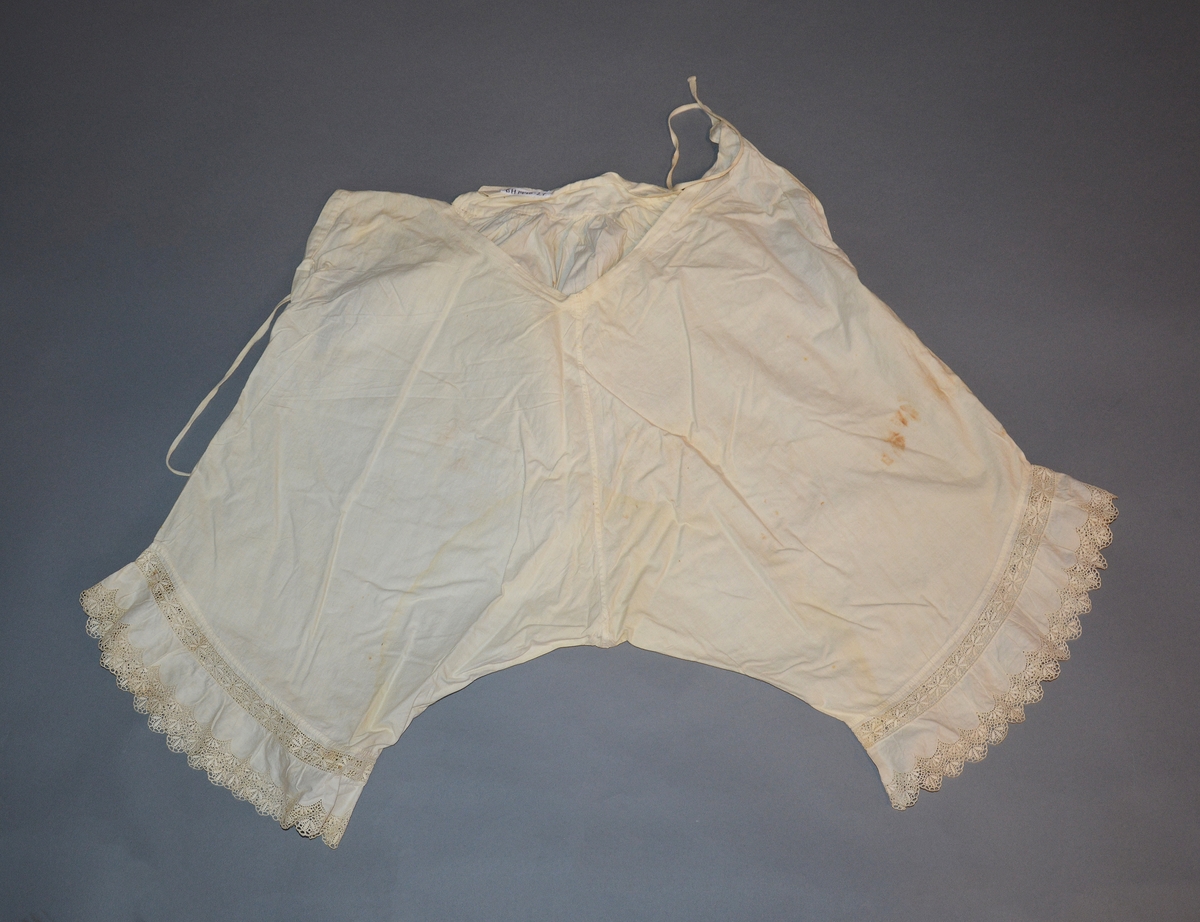 Underbukse for dame i bomullslerret. Åpning i sidene med knytebånd, knapp og knapphull. Bred linning bak, linning foran skåret ned i spiss og kantet med bendelbånd. Innfelt bomullsblonde nede på bena, og kantet med bomullsblonde.